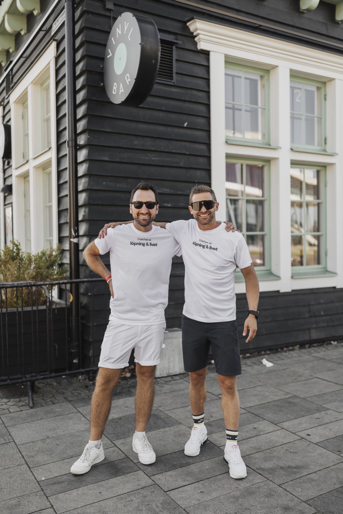Simon och Fredrik i vita löpning & livet-tröjor framför ett svart trähus